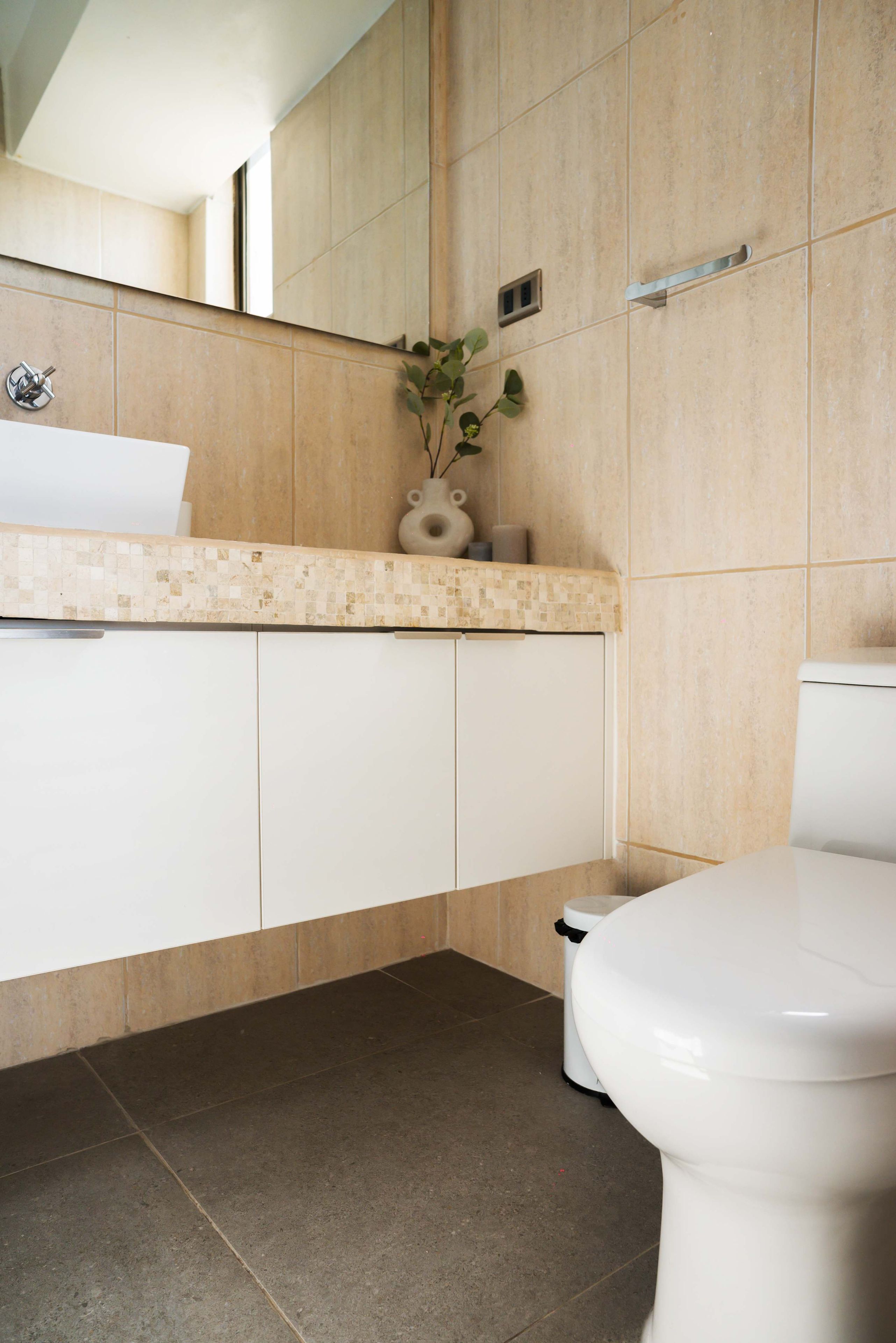 A bathroom with a white toilet, a beige tiled wall, and a white sink with a mosaic countertop.