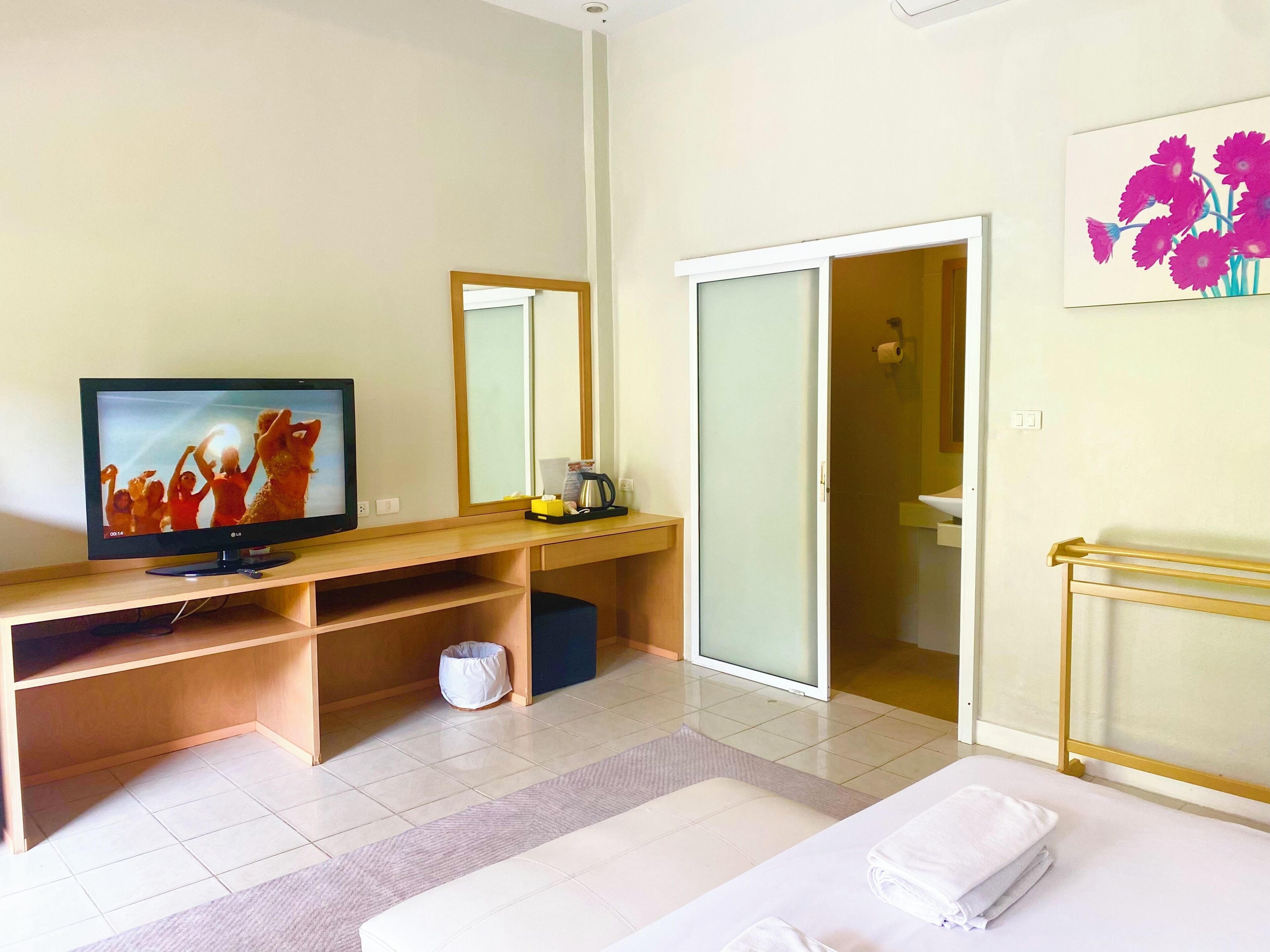 A hotel room with a TV, a desk with a coffee maker, a bed with towels, a mirror, and a painting of flowers.