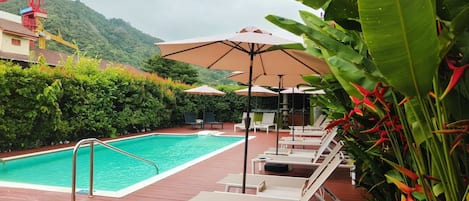 A swimming pool with lounge chairs and umbrellas, surrounded by greenery and a mountainous backdrop.