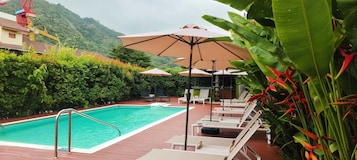 Piscina con sillas de descanso y sombrillas, rodeada de vegetación y montañas al fondo.