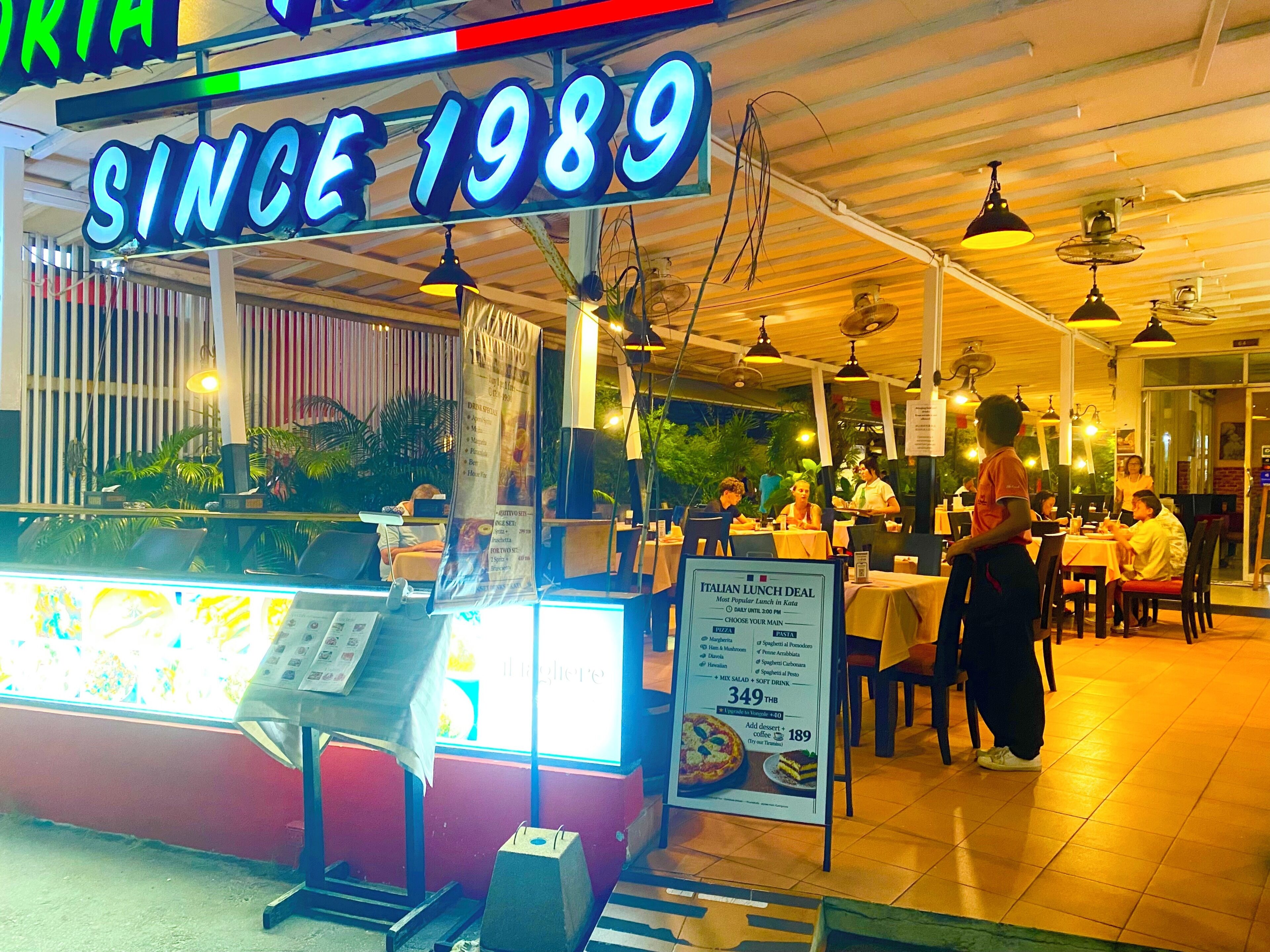 Un área de comedor al aire libre con un letrero de neón que dice 'SINCE 1989' y un panel publicitario con ofertas de comida italiana para el almuerzo.