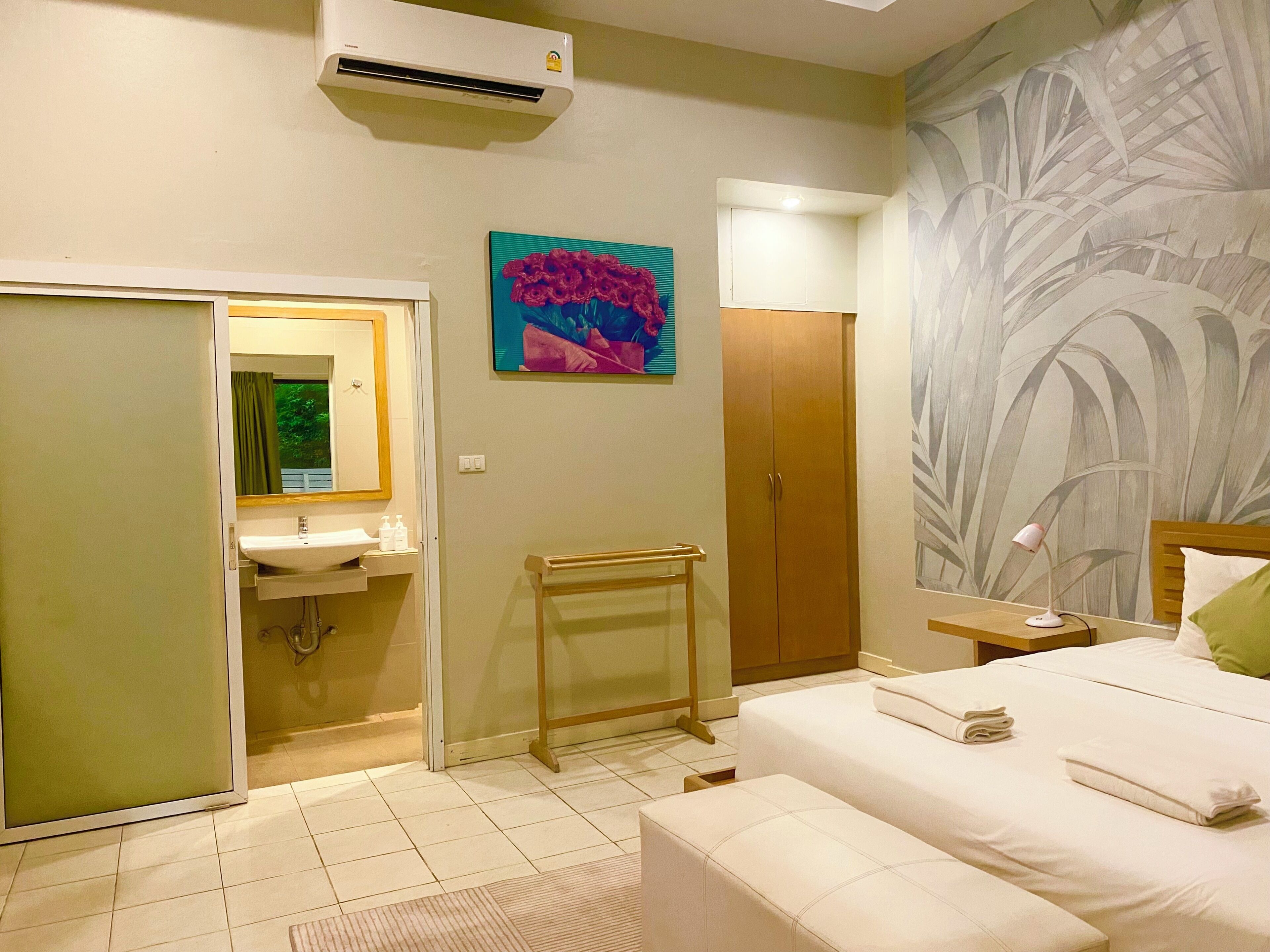 A hotel room with a bed, a bedside table, a nightstand, a wall-mounted air conditioner, a bathroom with a sink and mirror, and a towel rack.