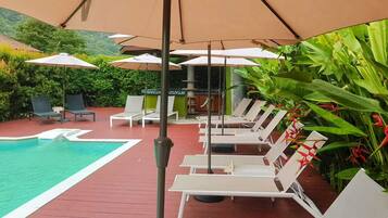 Zona de piscina con sillones reclinables y sombrillas, rodeada de follaje.