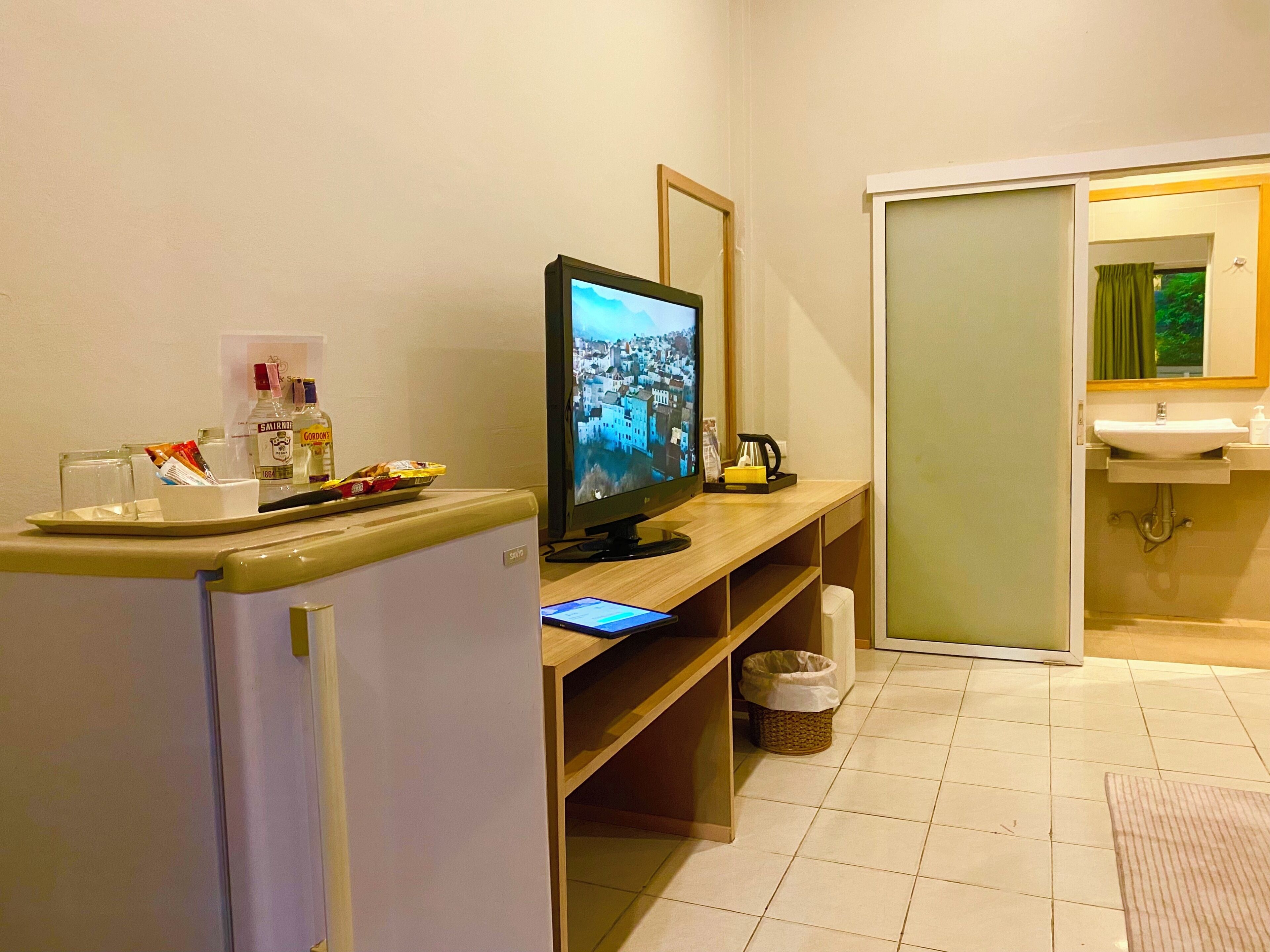 Hotelkamer met televisie, kleine koelkast, dienblad met glazen en snacks, mand, en een badkamer met wastafel en spiegel.