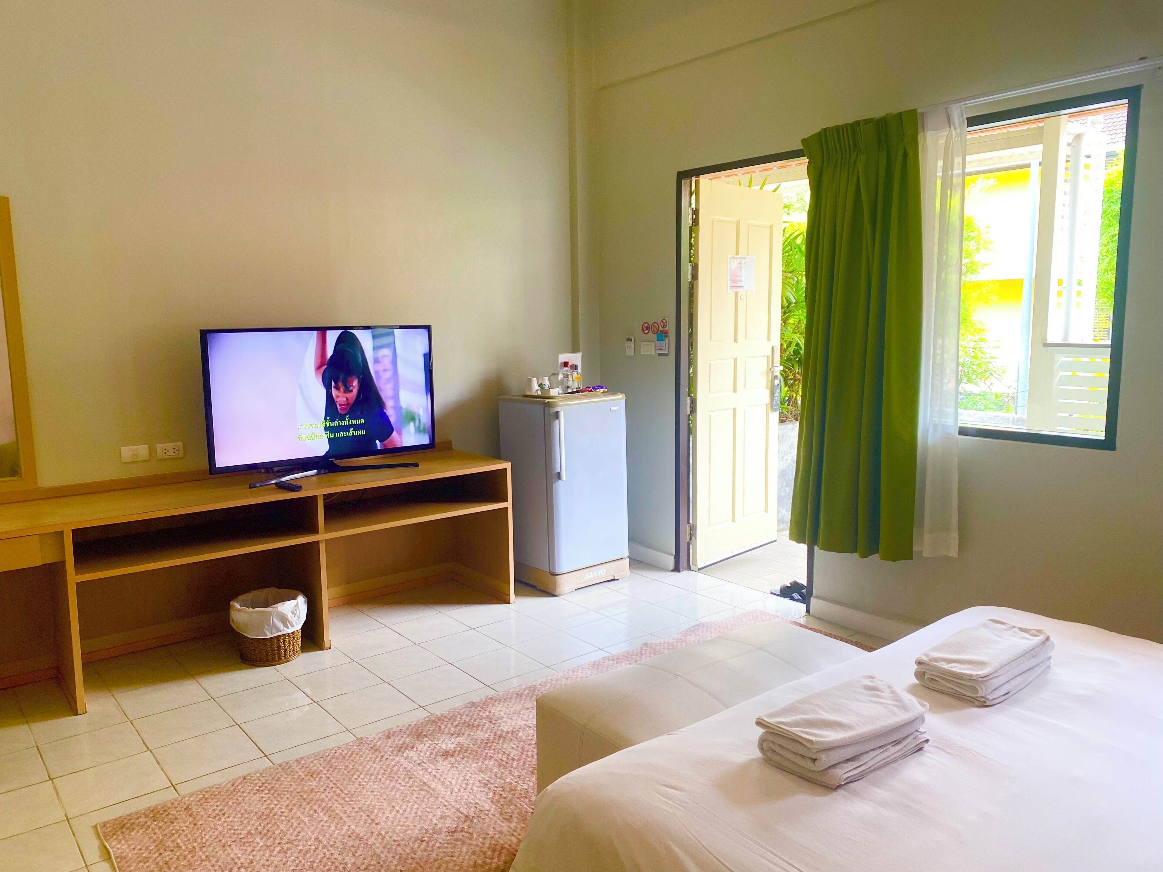 A hotel room with a television, a refrigerator, a bed with white linens, a small table, and a window with curtains.