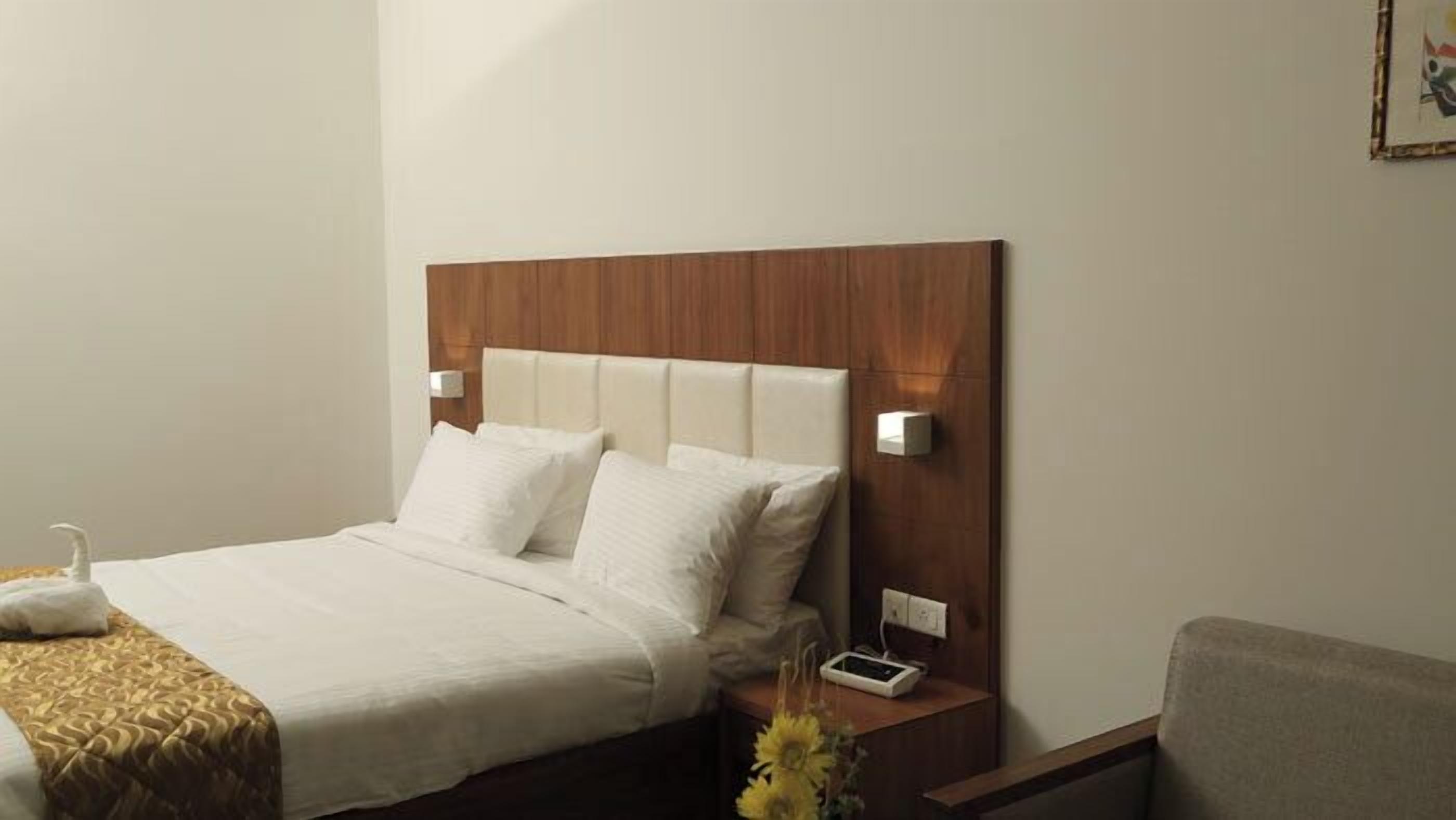 A neatly made bed with white linens, a wooden headboard, bedside lamps, and a bedside table with a phone and a vase of flowers.