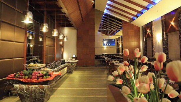 Un hall de hotel con un mostrador de recepción alargado, una pantalla de televisión y una zona de bufé con frutas y flores.