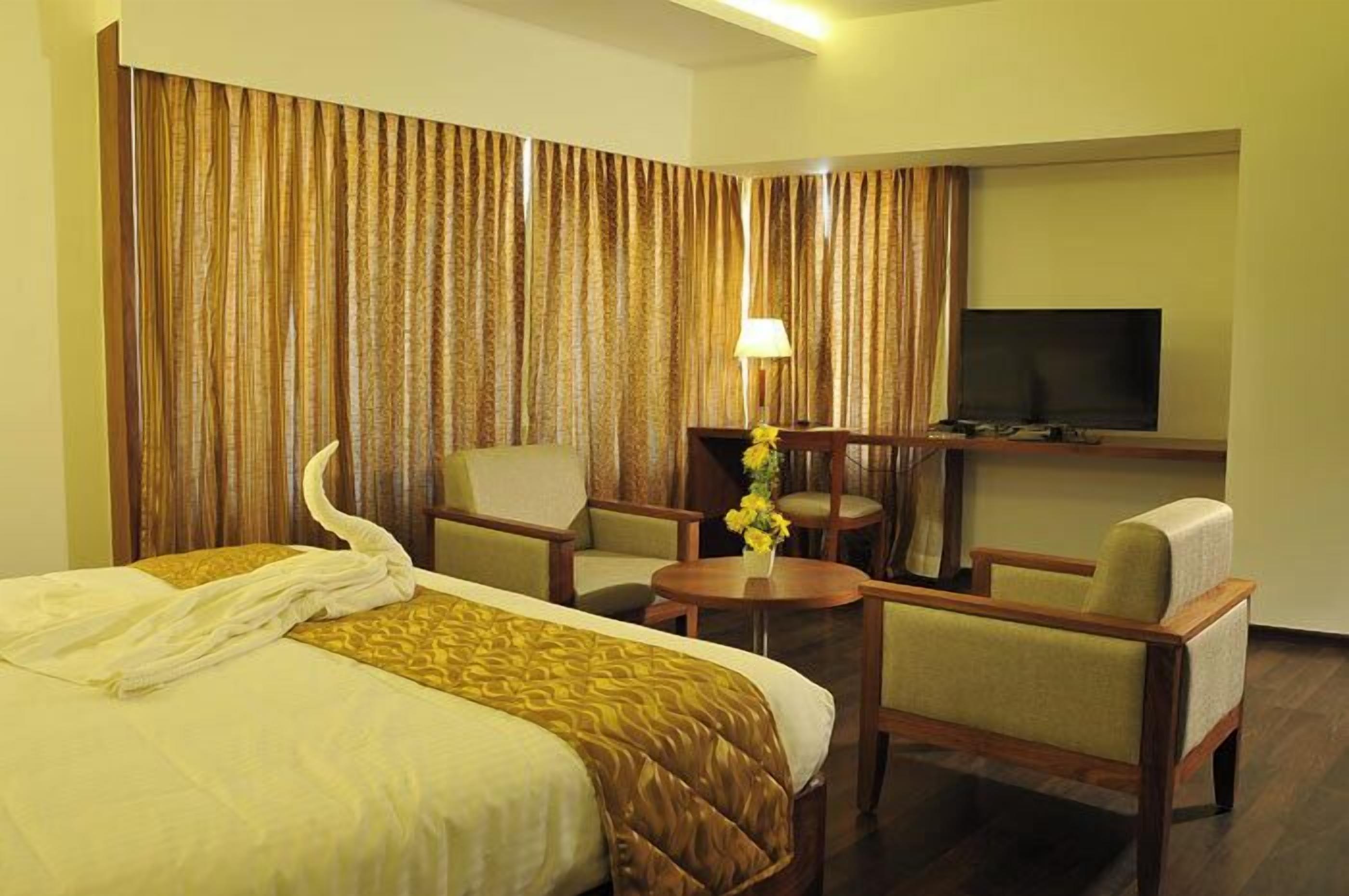 A hotel room with a bed, two armchairs, a small table with a lamp, and a TV on the wall.