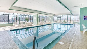 Une piscine intérieure avec des parois en verre transparent, un sol carrelé et un espace détente avec des chaises et des tables.