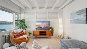 A modern living room with a large window, a ceiling fan, a wooden entertainment unit, and a view of the water outside. - Mission Beach Gem - 1BR Near the Ocean (San Diego)