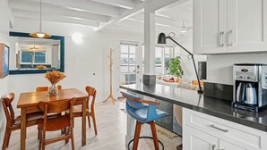 A modern kitchen with white cabinets, a coffee maker, and a dining area with wooden chairs and a vase of dried flowers. - Mission Beach Gem - 1BR Near the Ocean (San Diego)