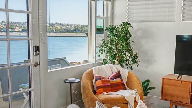 A cozy room with a large window overlooking a body of water, a comfortable armchair with a blanket, a small side table, and a TV stand. - Mission Beach Gem - 1BR Near the Ocean (San Diego)