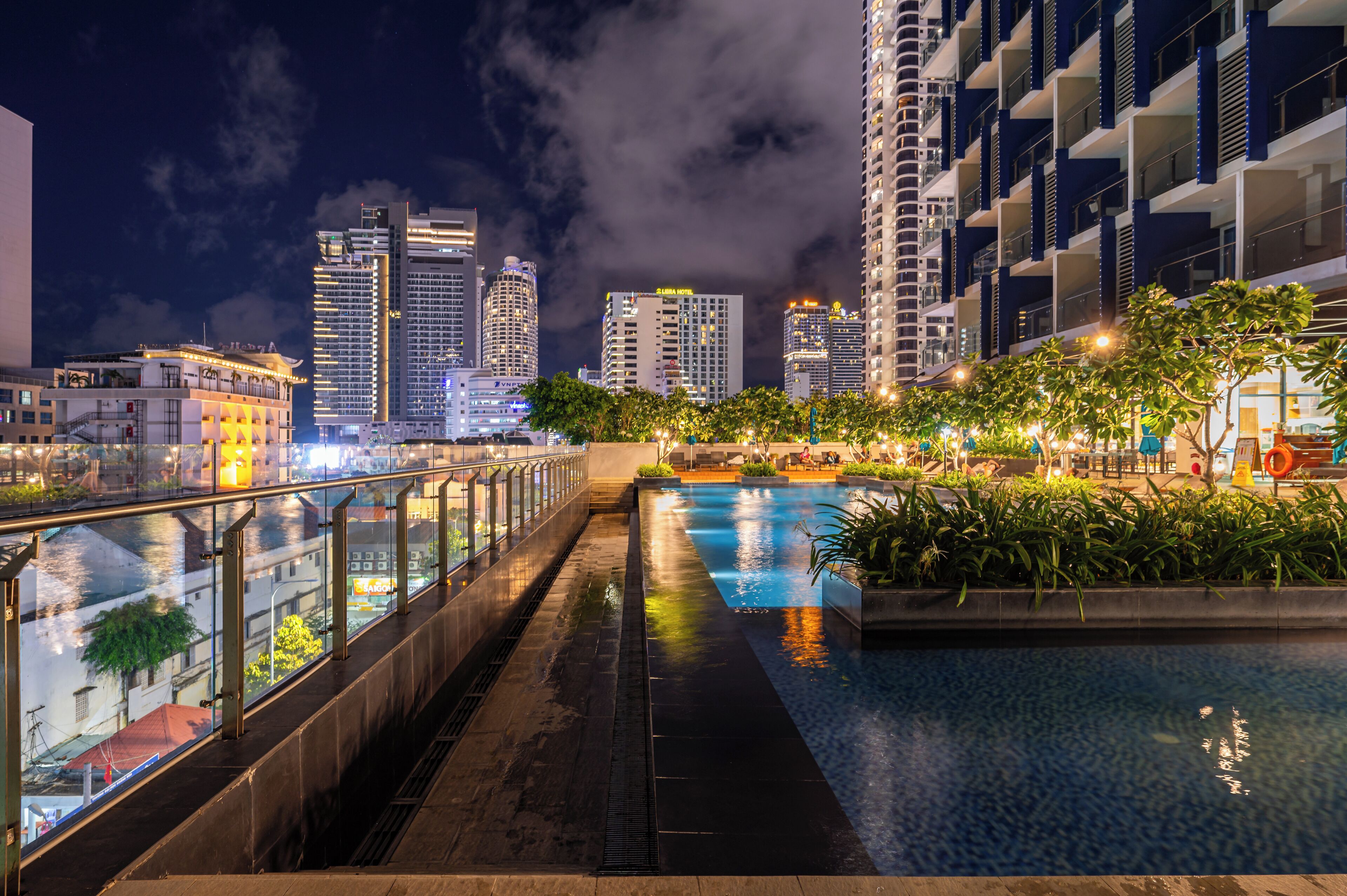 A nighttime view of a modern hotel with a pool area, featuring a glass railing, a water feature, and a city skyline in the background.
