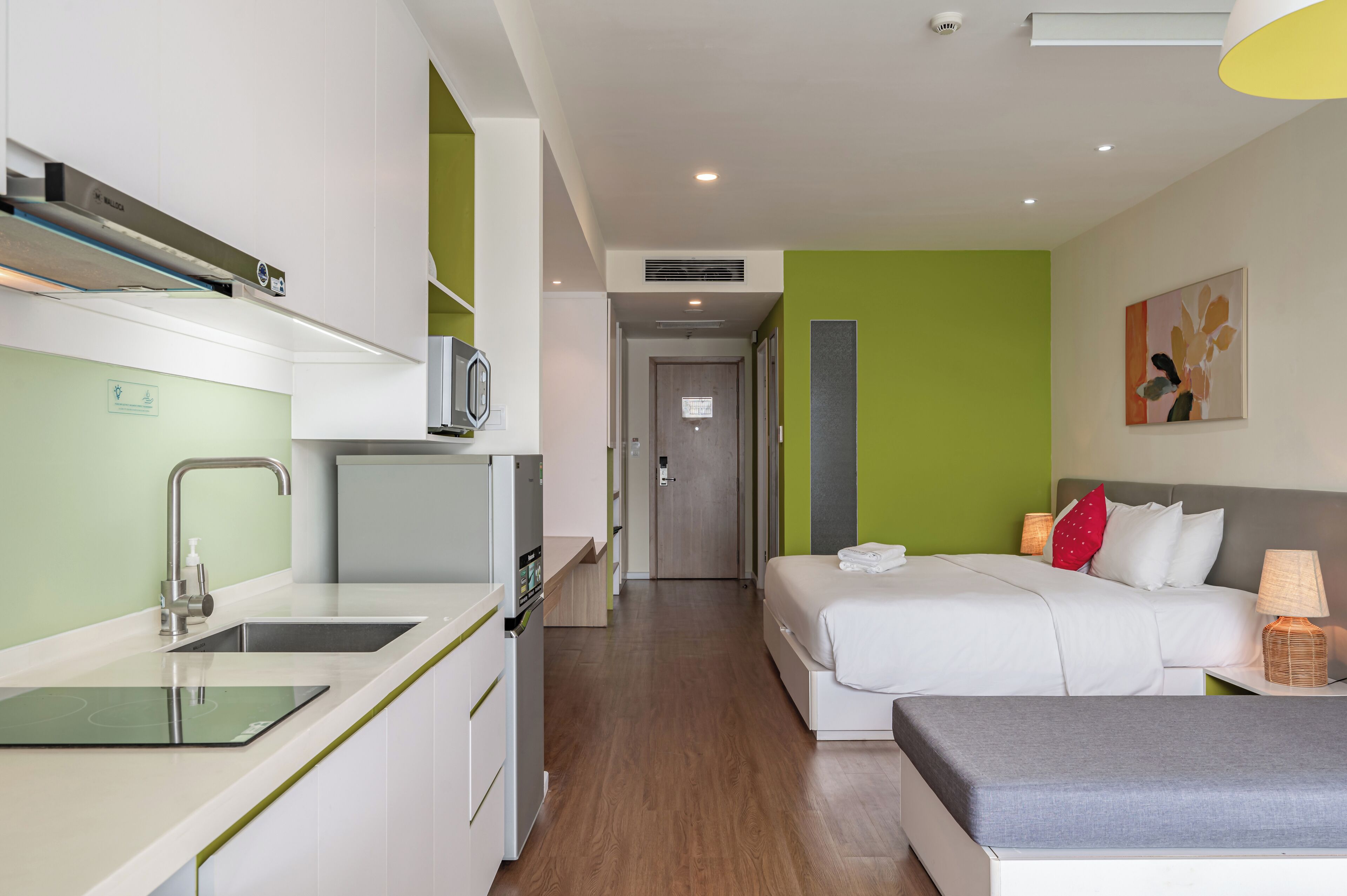 Une chambre d’hôtel moderne dotée d’une kitchenette, d’un lit avec une literie blanche, d’une table de chevet avec une lampe et d’un mur d’accent vert.