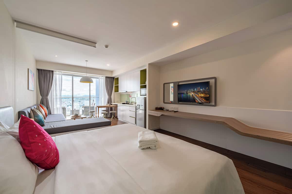 A hotel room with a large bed, a TV mounted on the wall, a desk with a chair, and a view of the city through the window.