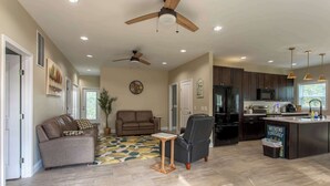 A spacious living area with a sofa, armchair, and a small wooden table. A ceiling fan and recessed lighting are visible.