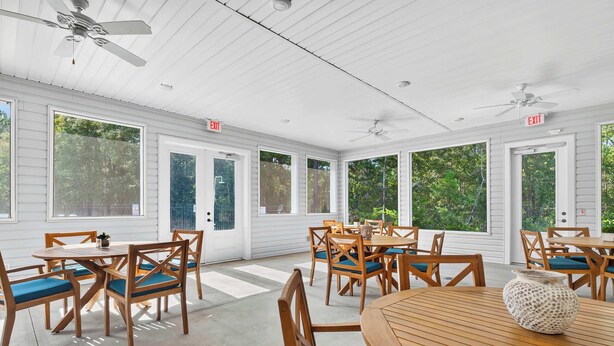 A covered patio area with wooden tables and chairs, large windows, and ceiling fans.