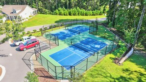 An outdoor tennis court surrounded by a fence, with a house and parked cars in the background.