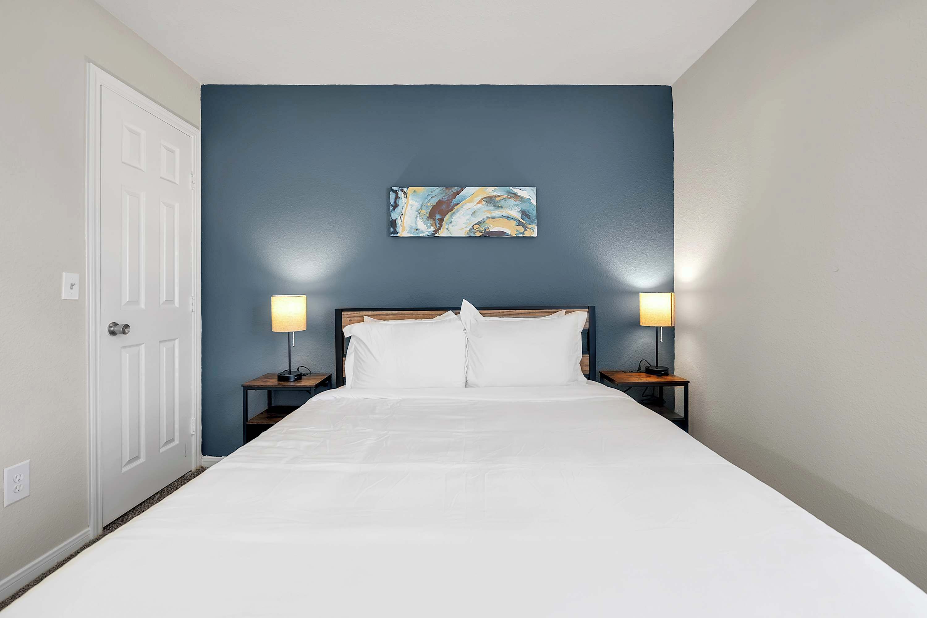 A neatly made bed with white linens, two bedside lamps, and a painting on the wall.