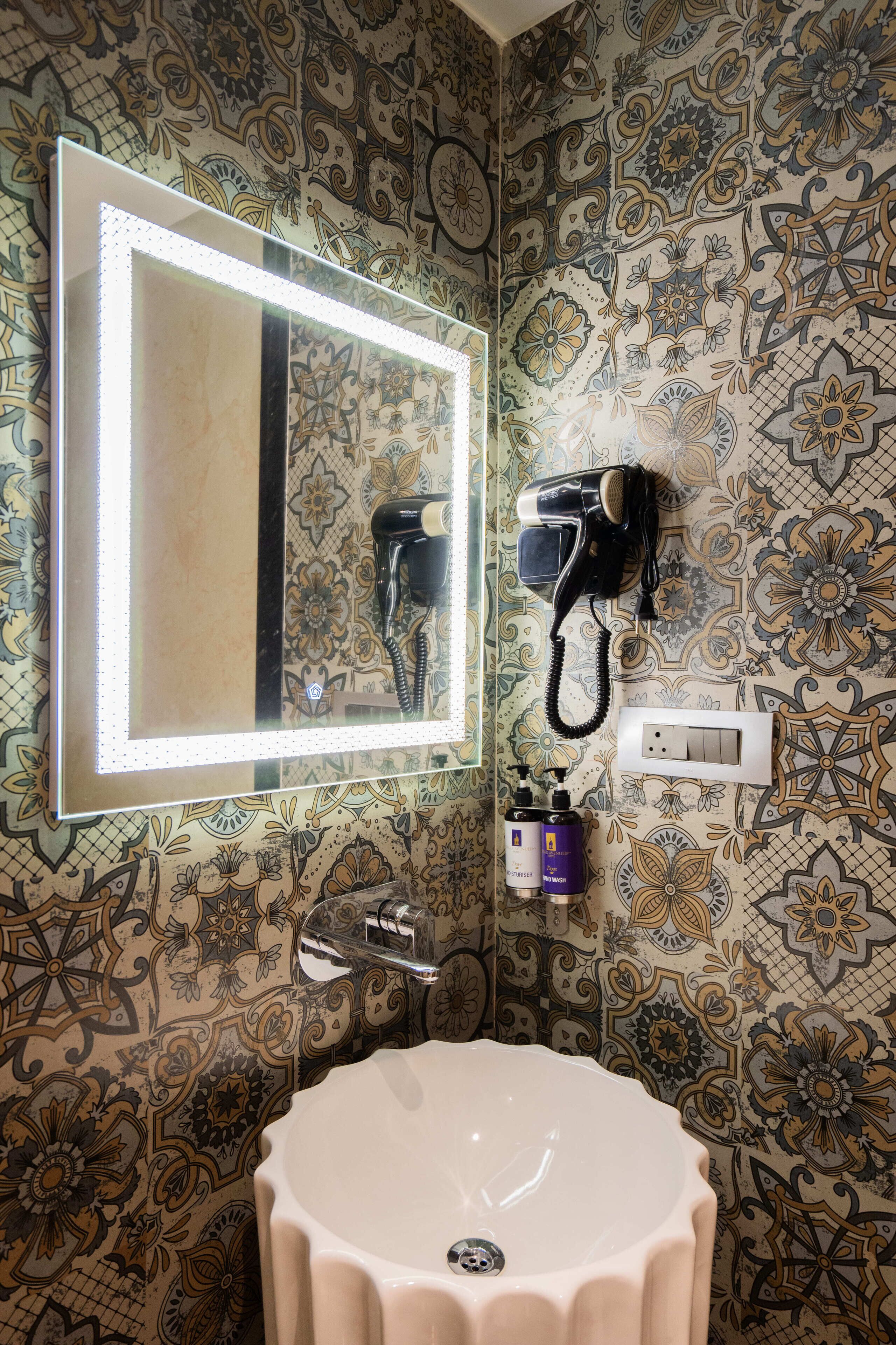Un bagno con un grande specchio, un asciugacapelli montato a parete, un lavandino e carta da parati decorata.