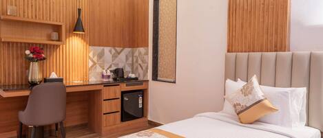Una habitación de hotel con un escritorio de madera, una cama con colcha estampada y un refrigerador pequeño.