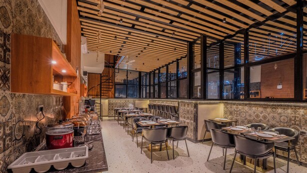Un restaurante con techo de madera, paredes enlosadas y una zona de bar. Mesas y sillas dispuestas para comer.