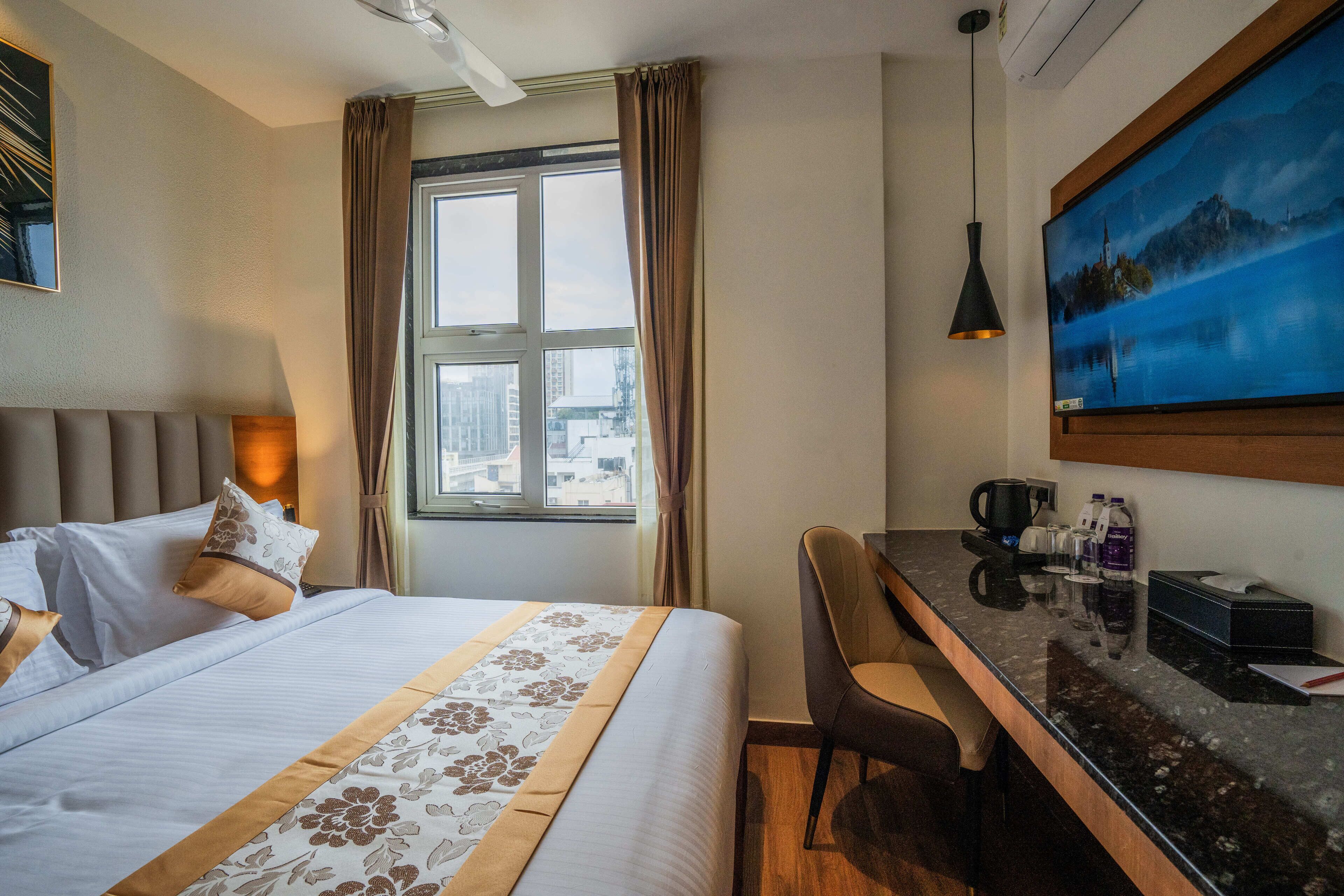 A hotel room with a bed, a desk, a chair, a large window with curtains, a painting on the wall, and a coffee maker on the desk.