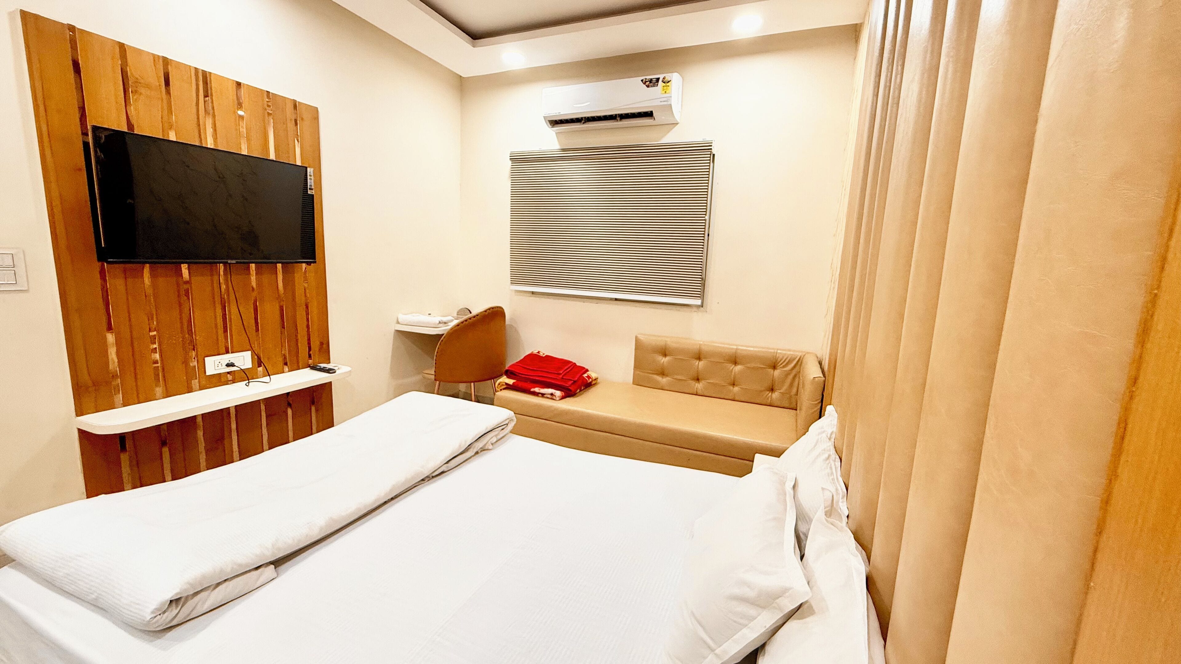 A hotel room with a bed, a TV mounted on a wooden panel wall, a window with blinds, a small desk with a chair, and a red towel on the bench.