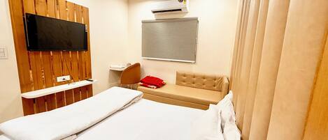 A hotel room with a bed, a TV mounted on a wooden panel wall, a window with blinds, a small desk with a chair, and a red towel on the bench.