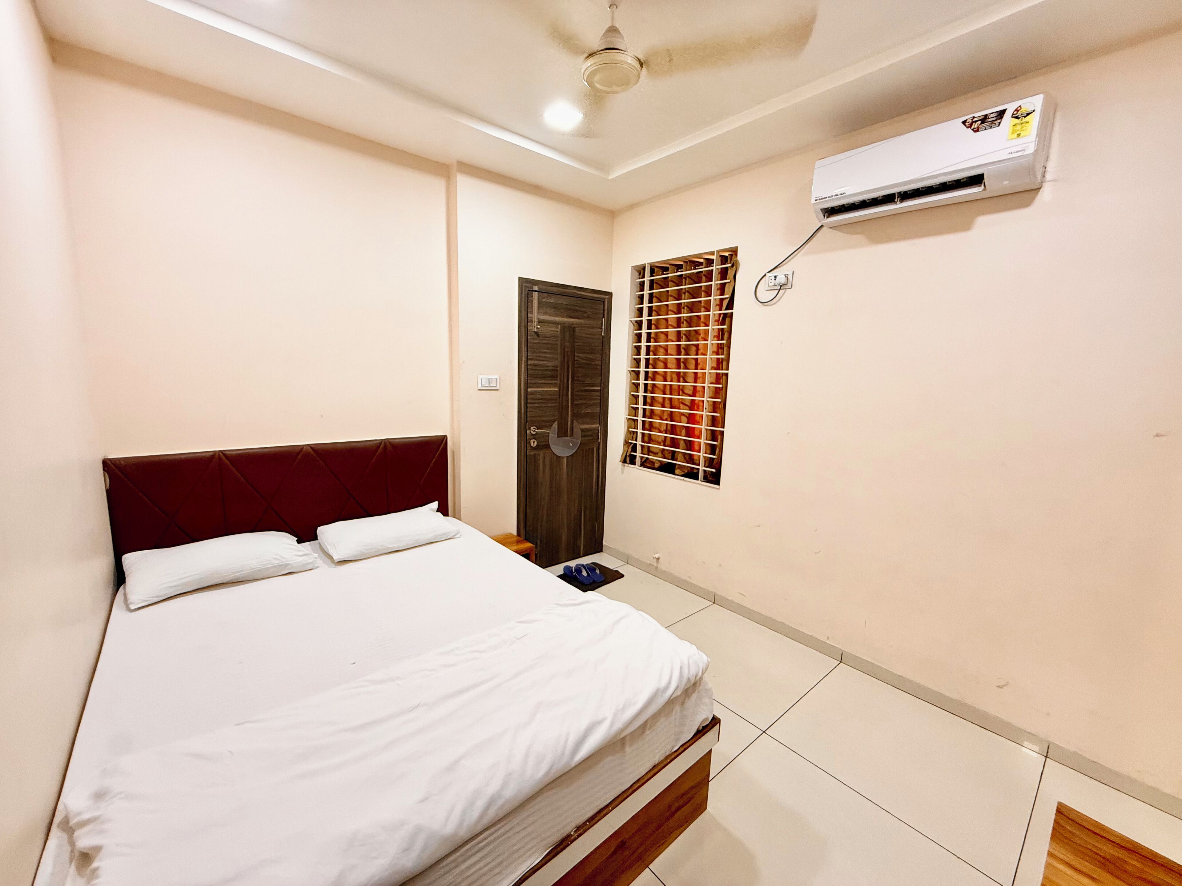 Une chambre à coucher comprenant un lit, un ventilateur de plafond, un appareil de climatisation, une fenêtre avec une grille en métal et une porte.
