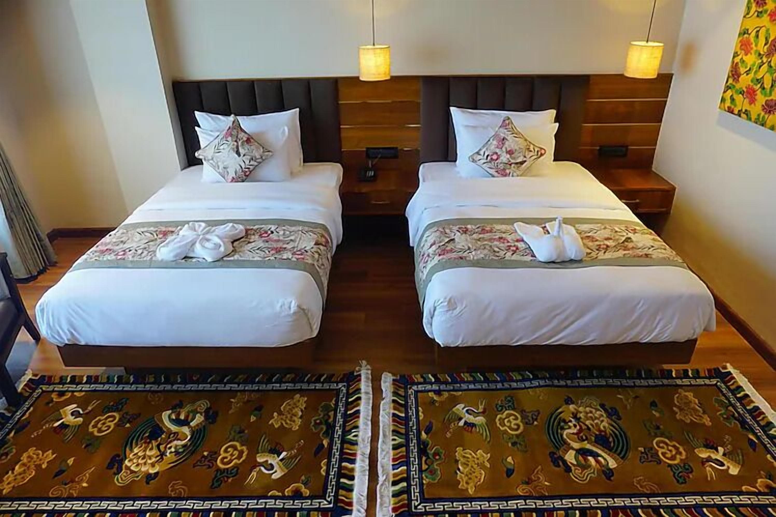 A hotel room with two beds, each with white linens and floral-patterned pillows. There are two bedside tables with lamps, and a decorative rug on the floor.