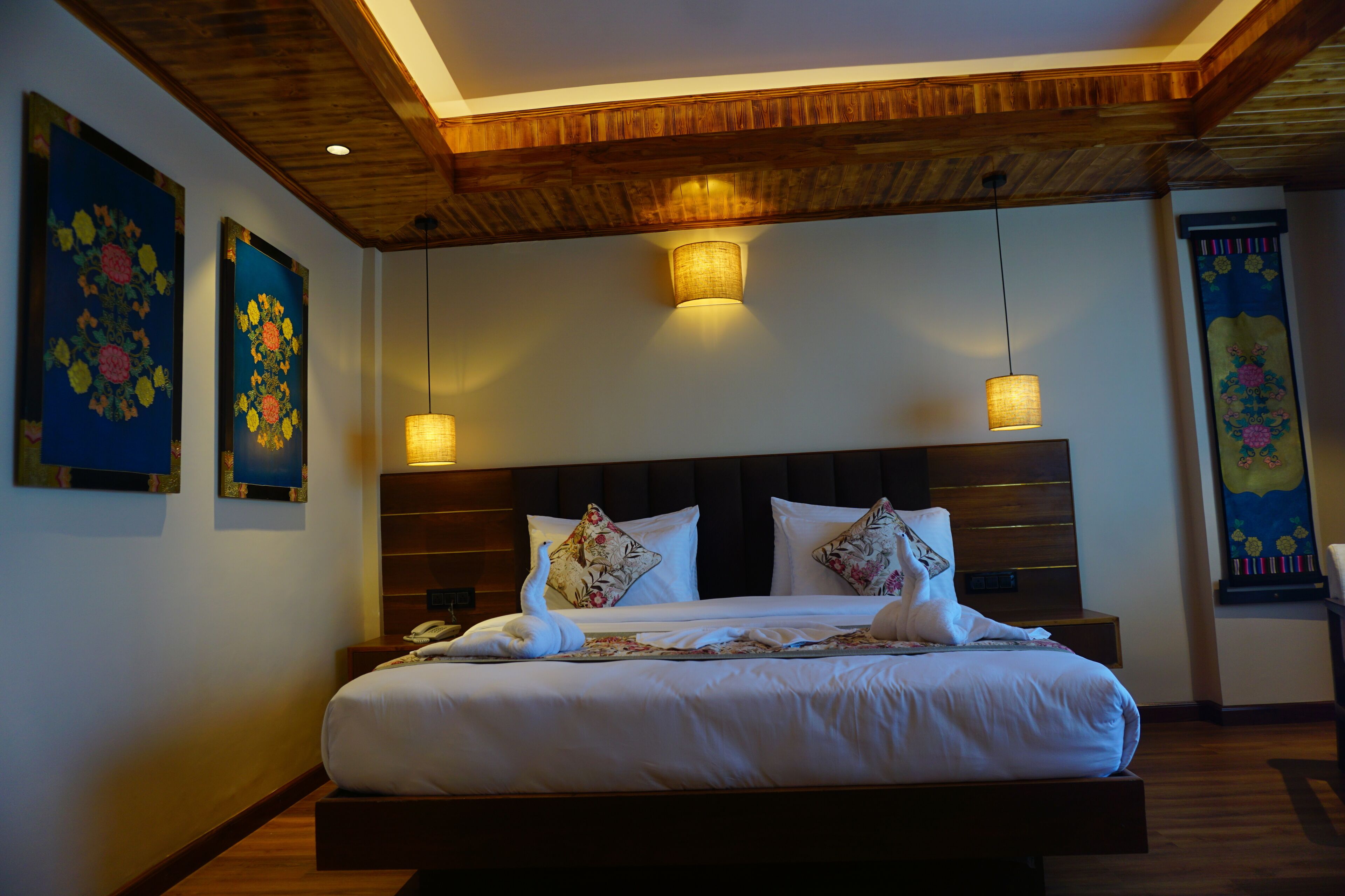 A hotel room with a wooden ceiling, a large bed with white bedding, two wall-mounted lamps, and floral artwork on the walls.