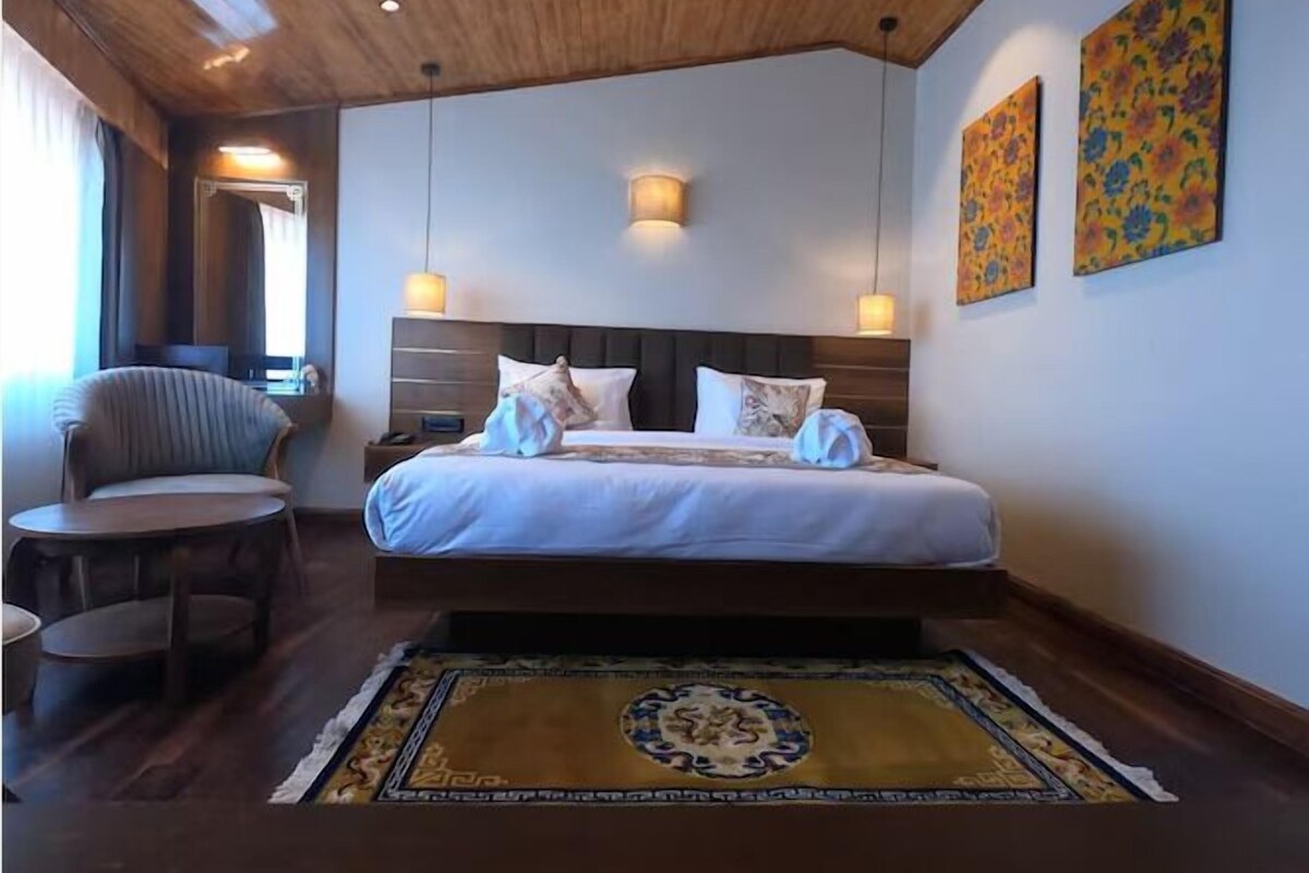 A hotel room with a wooden ceiling, a large bed with white bedding, two bedside lamps, a chair, and a decorative rug.