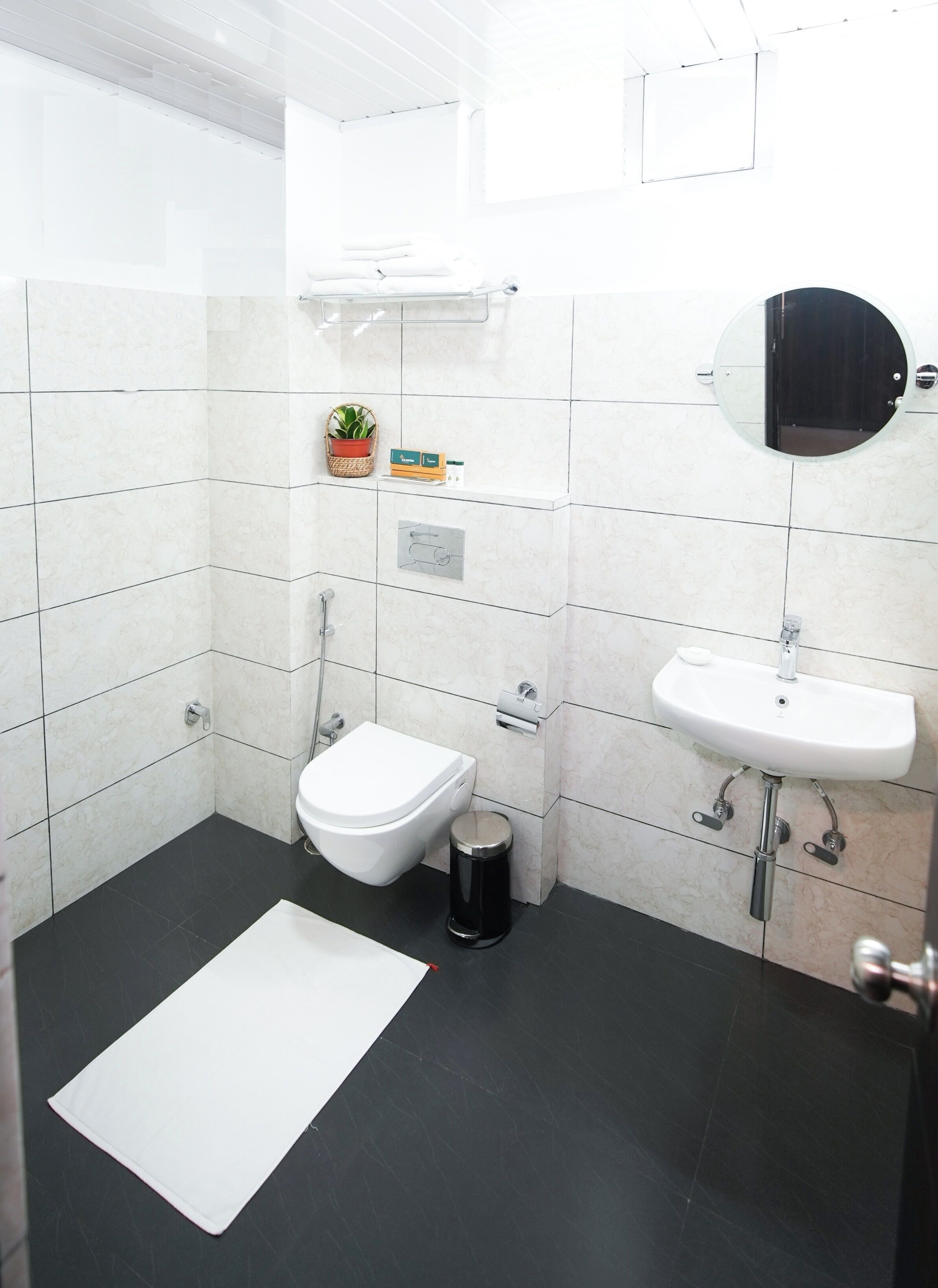 A modern bathroom with white tiles, a white toilet, a black trash bin, a white rug, and a round mirror above the sink.