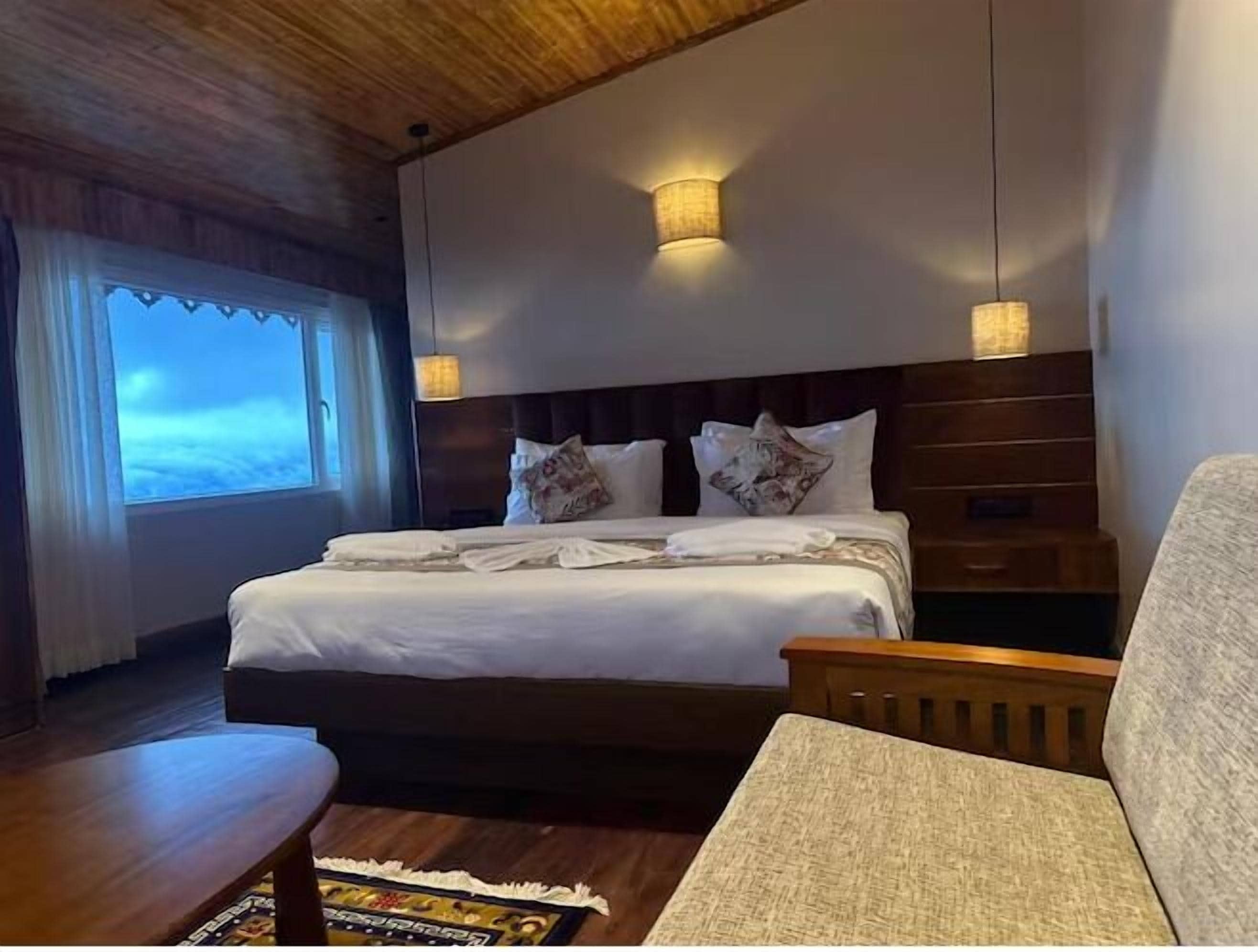 A hotel room with a large bed, two wall-mounted lights, a wooden headboard, a window with curtains, and a small table with a magazine.