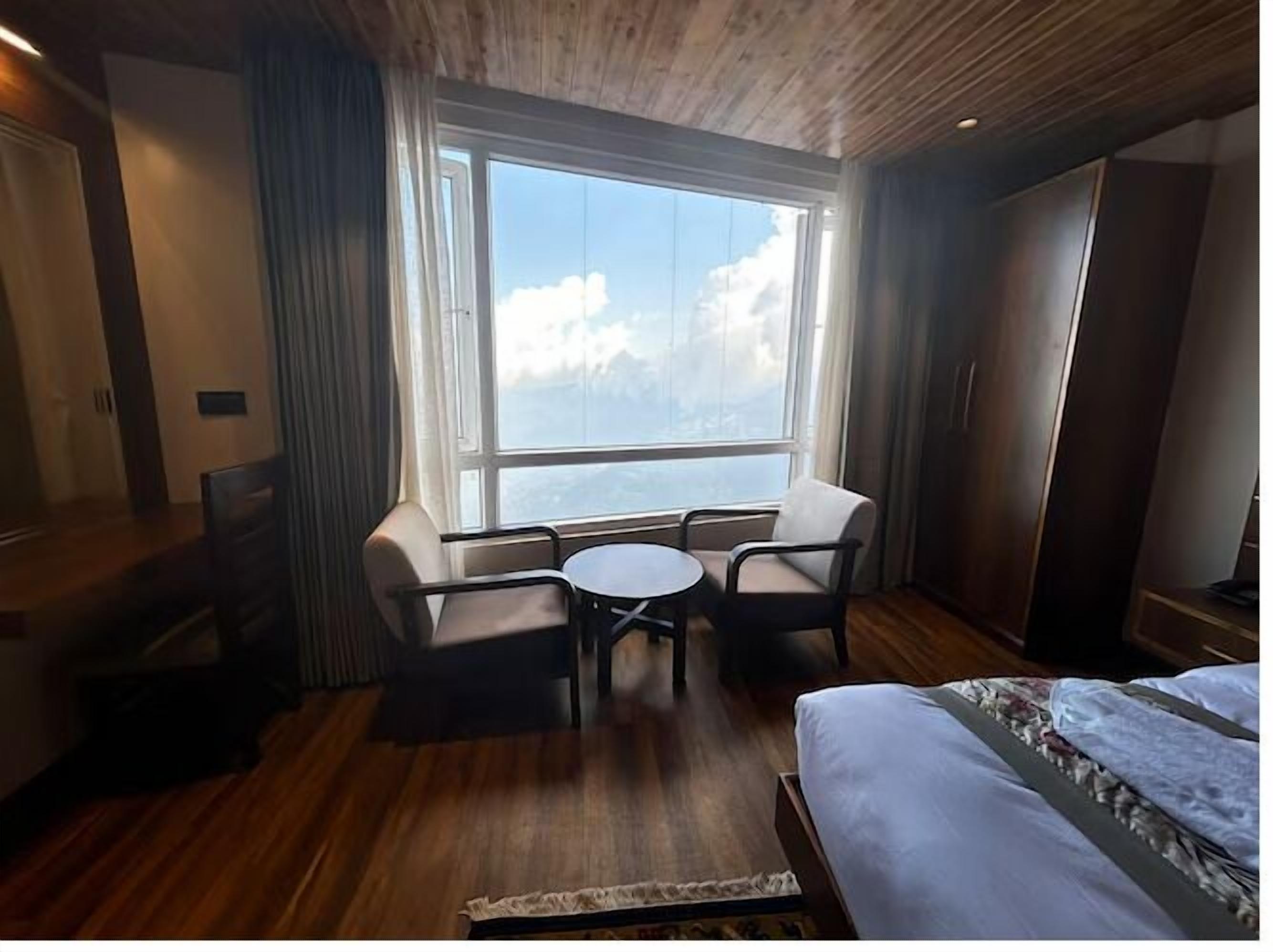 Una camera d'albergo con una grande finestra, un letto, un tavolino rotondo con due sedie e un pavimento in legno.