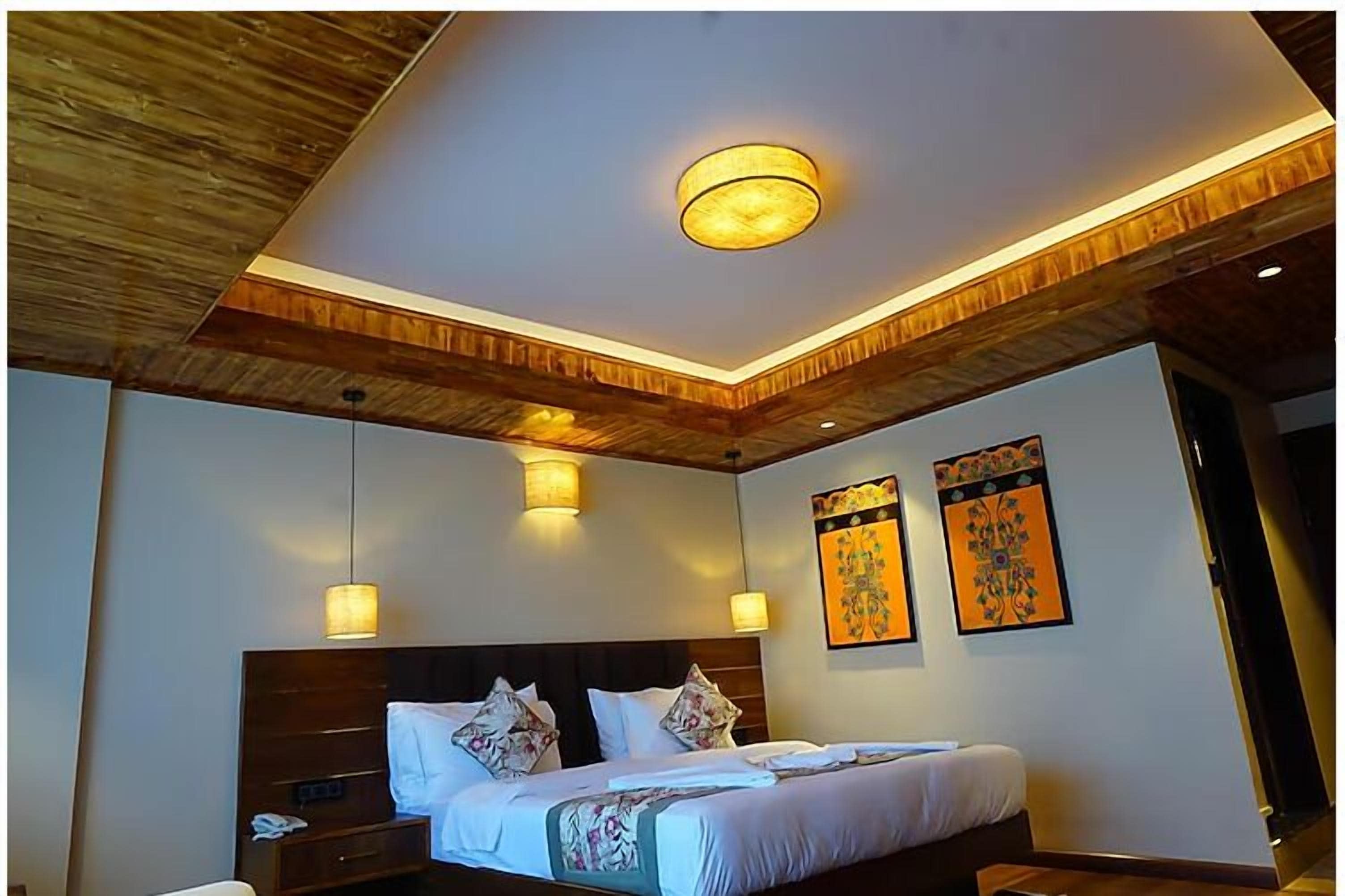 Habitación de hotel con techo de madera, una cama grande con ropa blanca, dos lámparas fijadas a la pared y dos cuadros con motivos florales.