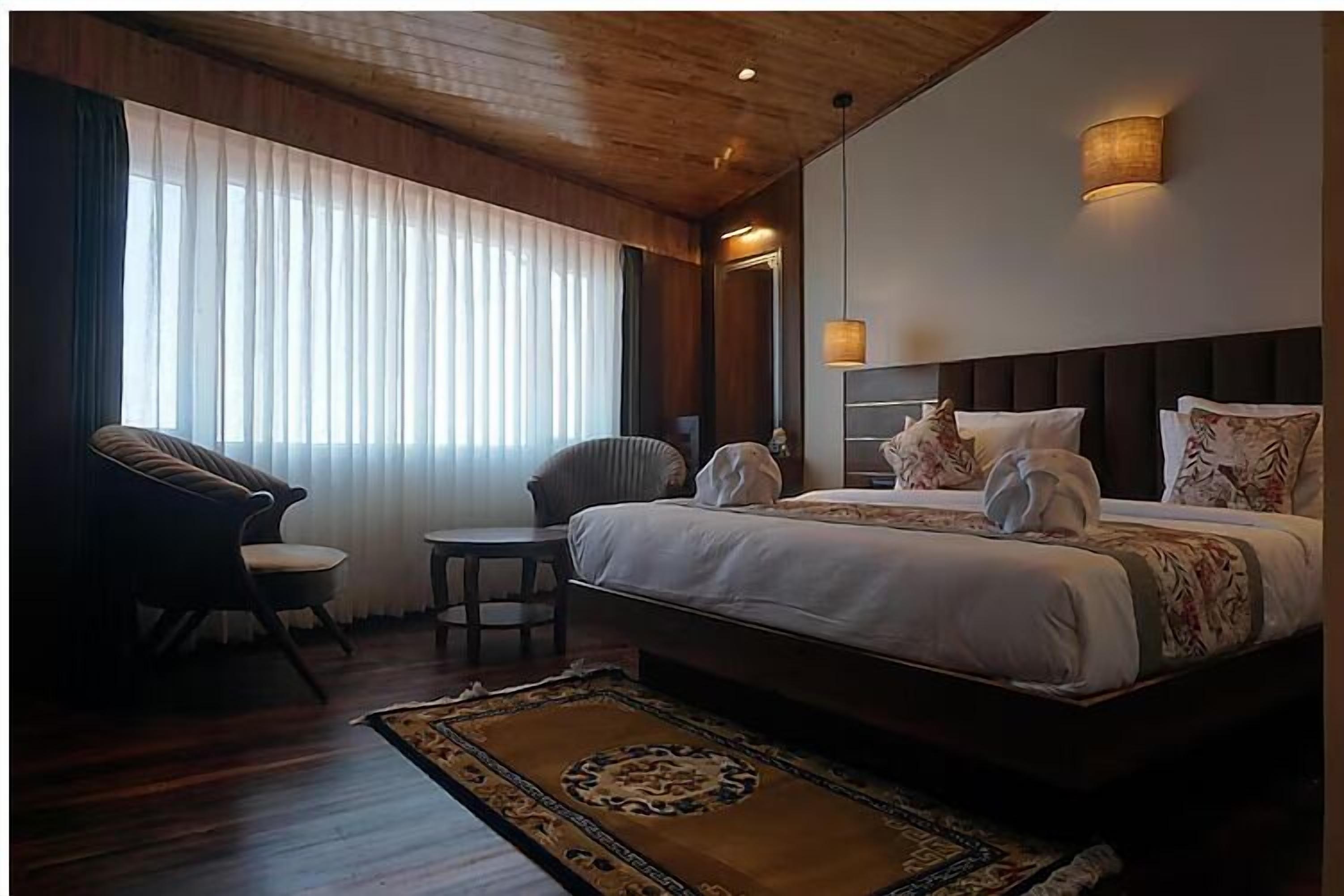 A hotel room with a large bed, two chairs, a small table, and a large window with sheer curtains.