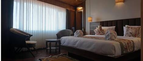 A hotel room with a large bed, two chairs, a small table, and a large window with sheer curtains.