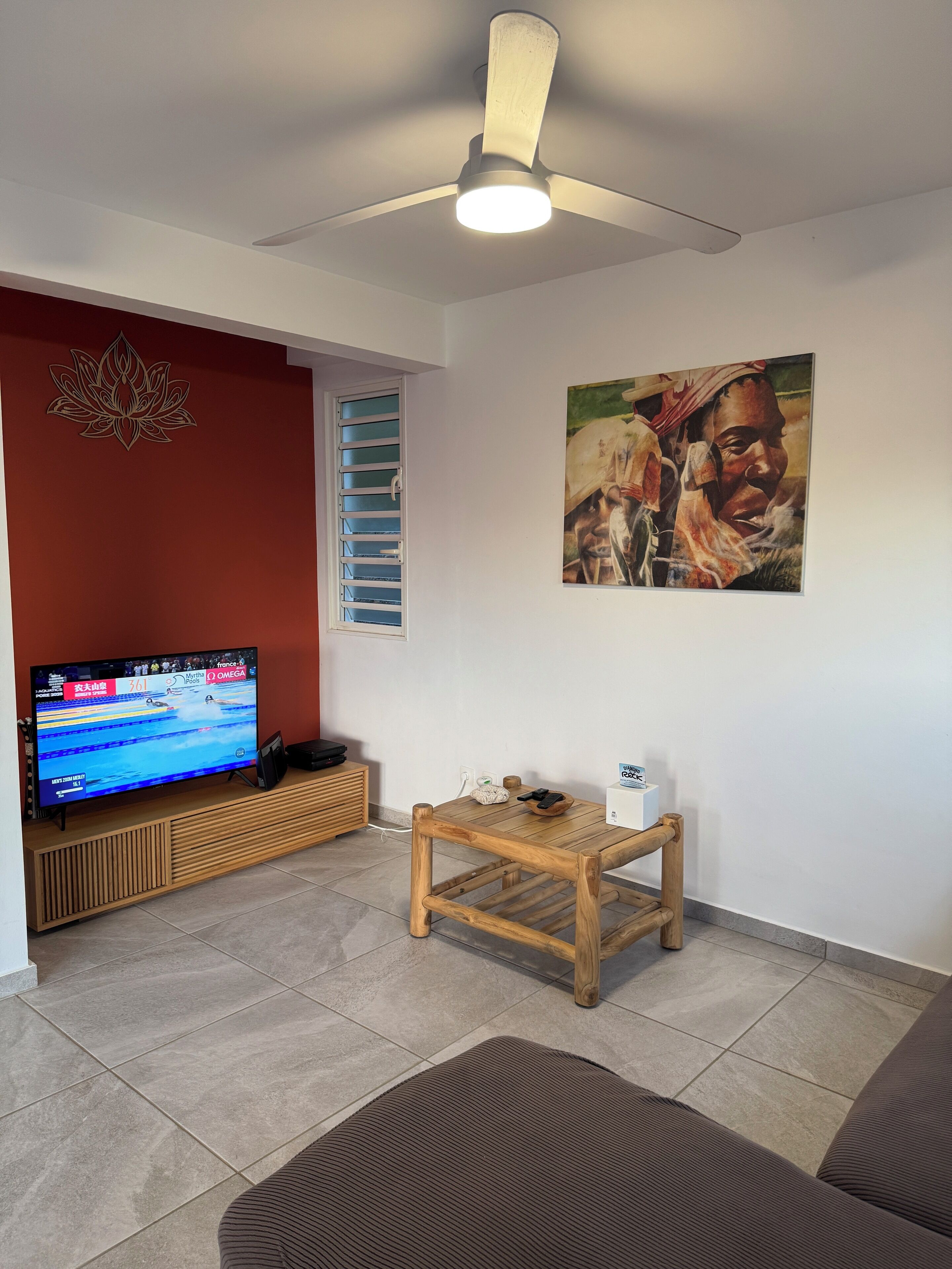 Una sala de estar con una televisión de pantalla plana, un ventilador de techo, una mesa de centro de madera, una pared de acento roja, una ventana con persianas y un cuadro en la pared.