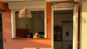 Un espace extérieur aménagé pour les repas, avec une table et des chaises, un luminaire suspendu, et une vue sur une cuisine équipée d’un réfrigérateur et de placards.