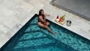 Une personne est assise au bord d’une petite piscine, dont le bord est recouvert de carreaux, et on aperçoit un bâtiment en arrière-plan.