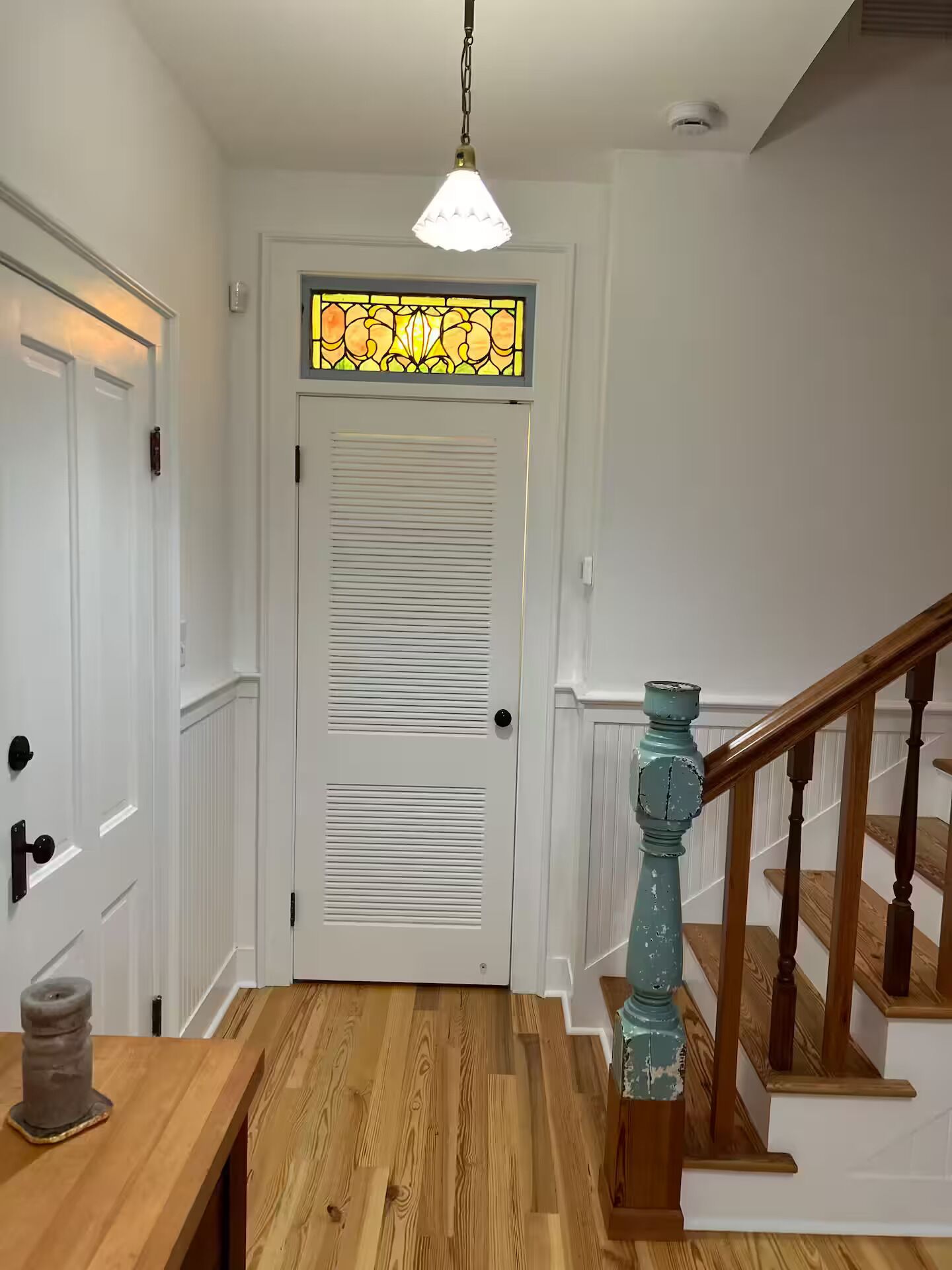 Un couloir doté d’un plancher en bois, de portes blanches, d’un vitrail au-dessus d’une porte et d’un escalier en bois avec une touche de couleur bleue.