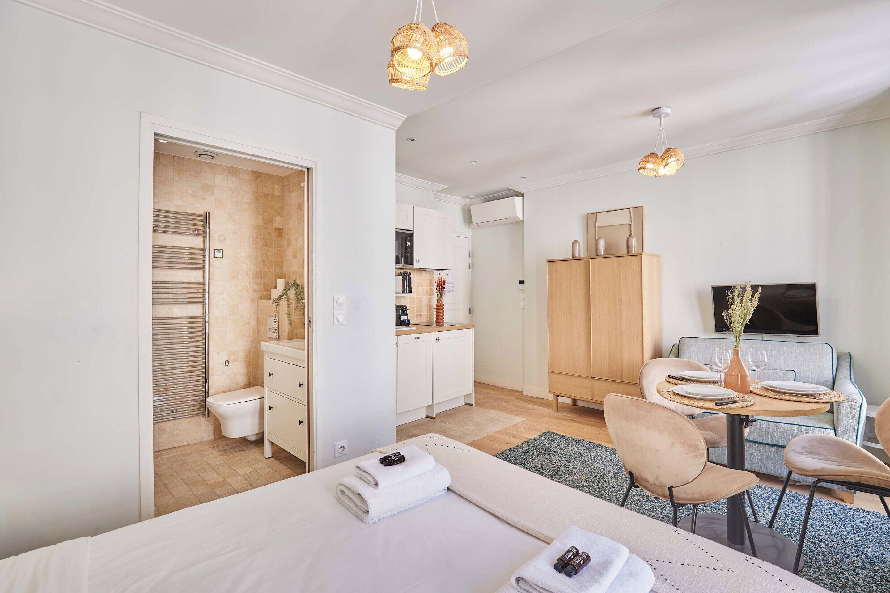 A modern hotel room with a bed, a dining area with a table and chairs, a kitchenette, and a TV.