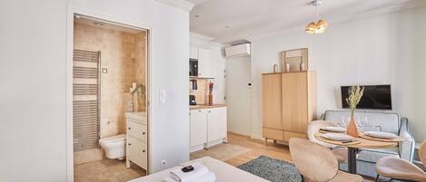 A modern hotel room with a bed, a dining area with a table and chairs, a kitchenette, and a TV.