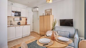 A modern kitchen with white cabinets, a microwave, and a coffee maker. There is a dining area with a round table, plates, and a vase with flowers. A sofa and a TV are also visible. - Superb Studio Center of Paris Montorgueil (Paris)