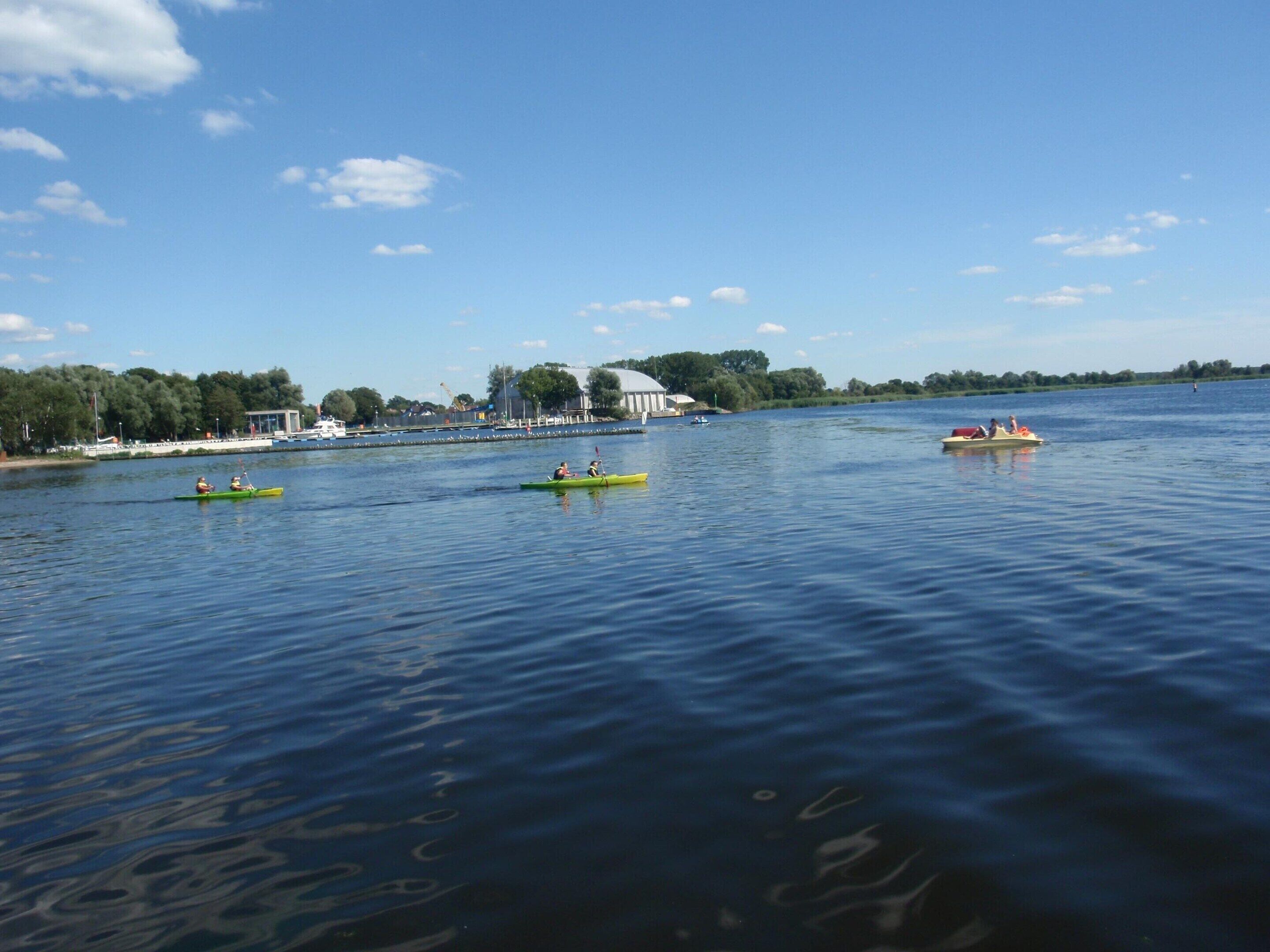 A calm lake with kayaks and a small boat. There are buildings and trees in the background.