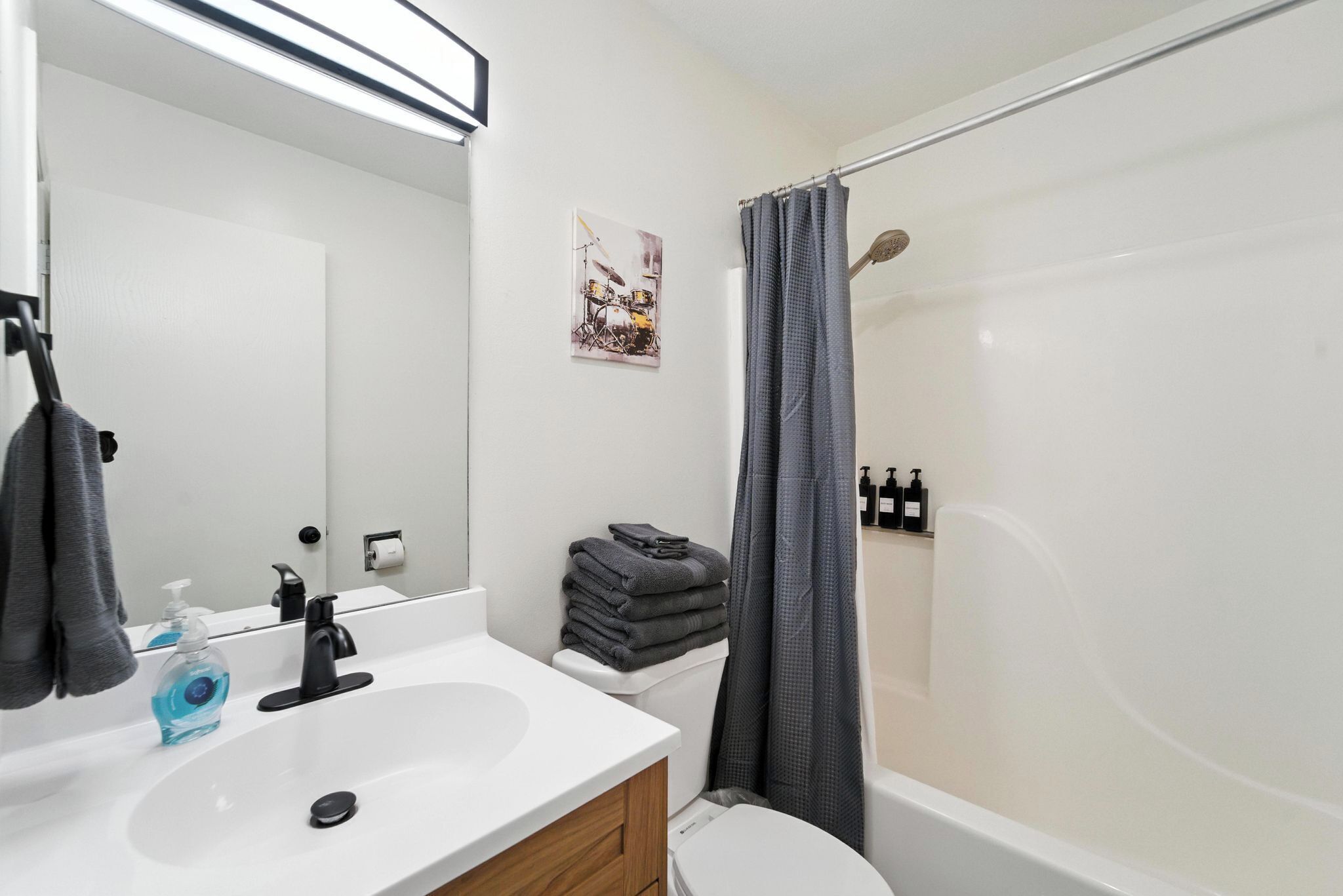 A modern bathroom with a white sink, black faucet, and a toilet. There is a shower curtain, a stack of towels, and a framed picture on the wall.