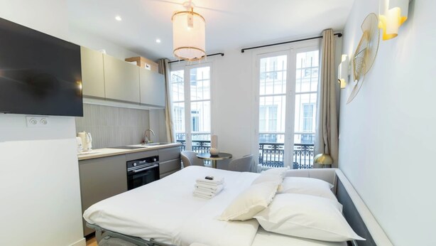 A modern hotel room with a bed, bedside table, and a view of the city through large windows. - Charming Studio - Le Marais - Bastille (Paris)