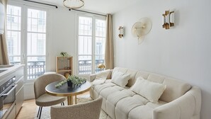 A modern living room with a beige sofa, a round dining table with two chairs, a small side table with a plant, and a large window with curtains. - Charming Studio - Le Marais - Bastille (Paris)